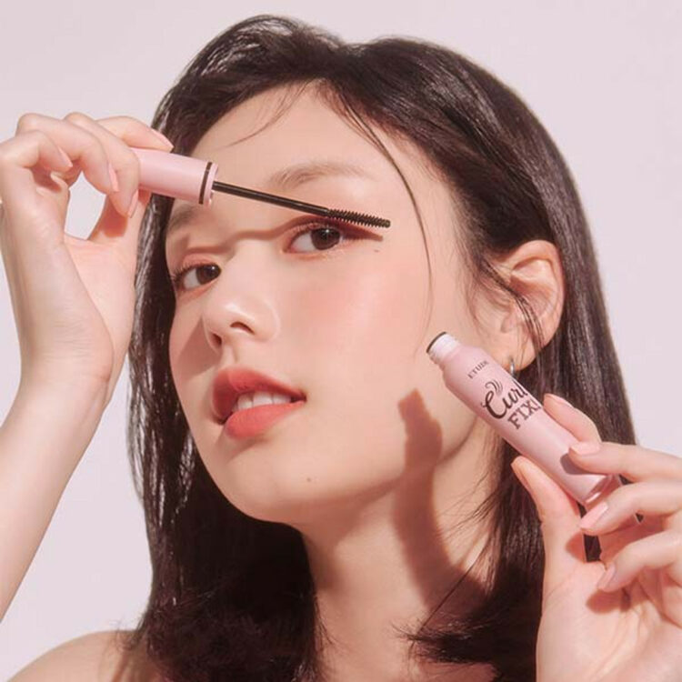 woman applying ETUDE Curl Fix Mascara. The tube is pink and the background is pink