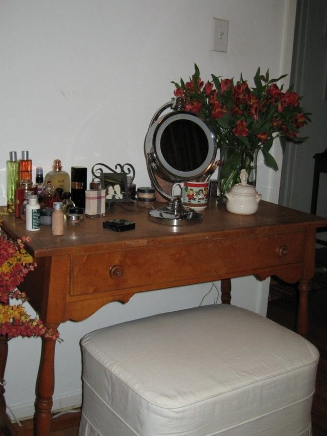 photo of the top of a dressing table with bottles of perfume and cosmetics, a vase with alstromeria flowers and a lighted makeup mirror.