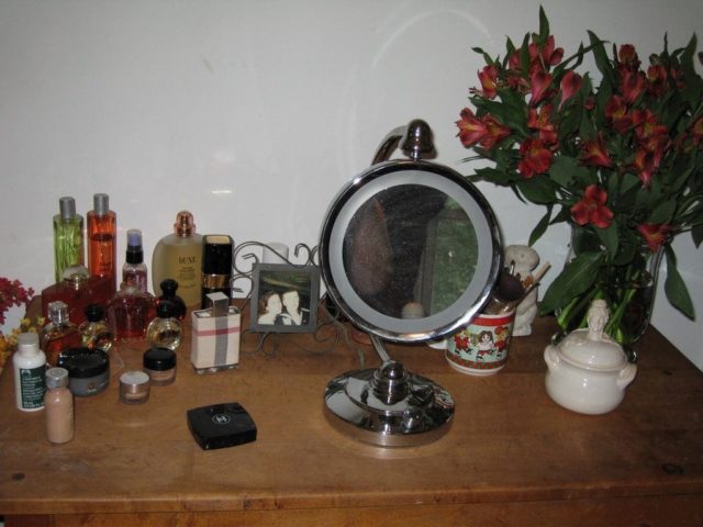photo of the top of a dressing table with bottles of perfume and cosmetics, a vase with alstromeria flowers and a lighted makeup mirror.
