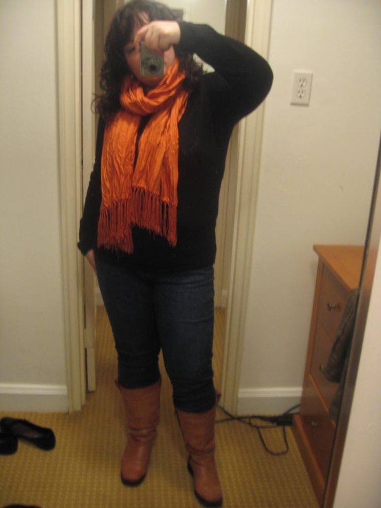 woman in a black sweater, jeans, tall tan boots, and a long orange fringed silk scarf around her throat