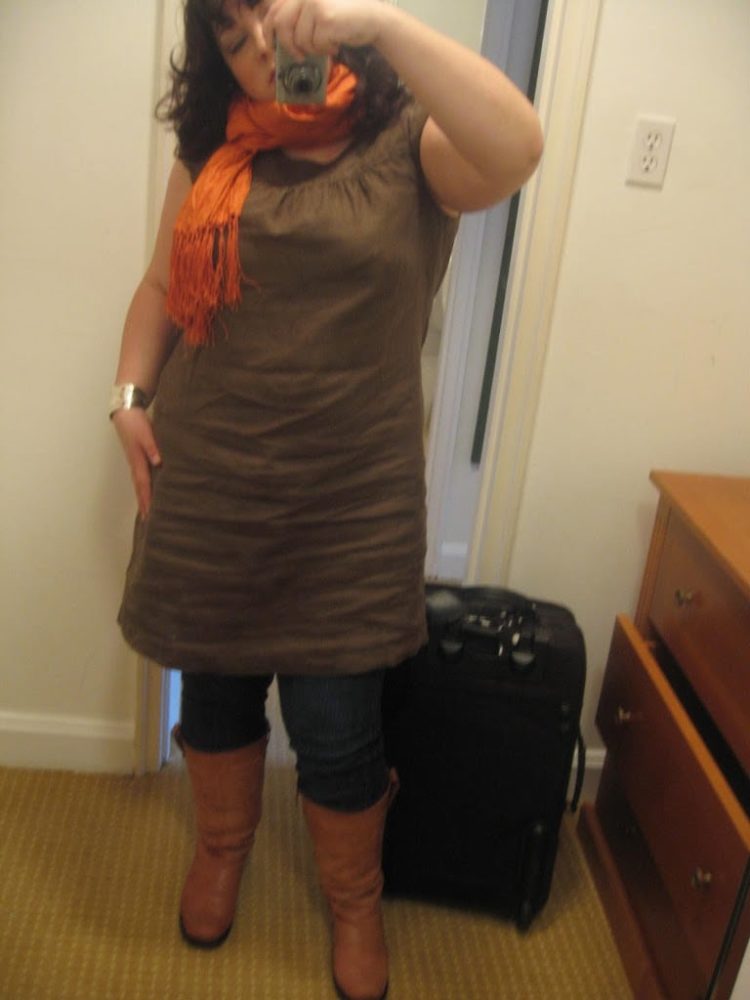 woman in an olive green linen shift over jeans and tall brown boots, an orange crinkle silk scarf looped around her neck