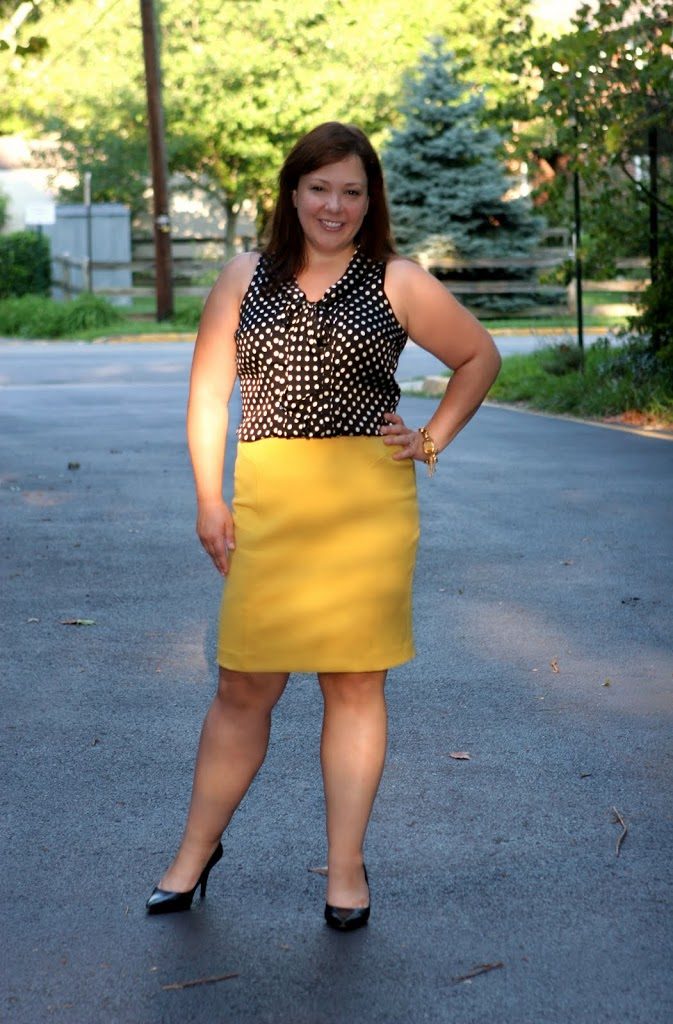 woman standing outside with her hand on her hip, smiling at the camera. She is wearing a black sleeveless blouse with white polka dots and it is tucked into a yellow pencil skirt. She is wearing black pumps and has shoulder length straight brown hair.