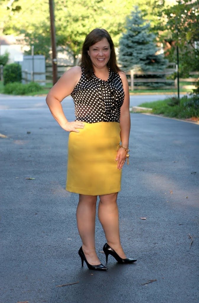 Woman in a sleeveless black and white polka dot blouse with bow detail at the neck an d a yellow pencil skirt