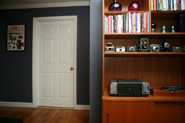 View of a white door against dark purple gray painted walls with a view of a mid century modern Scandanavian wood cabinet