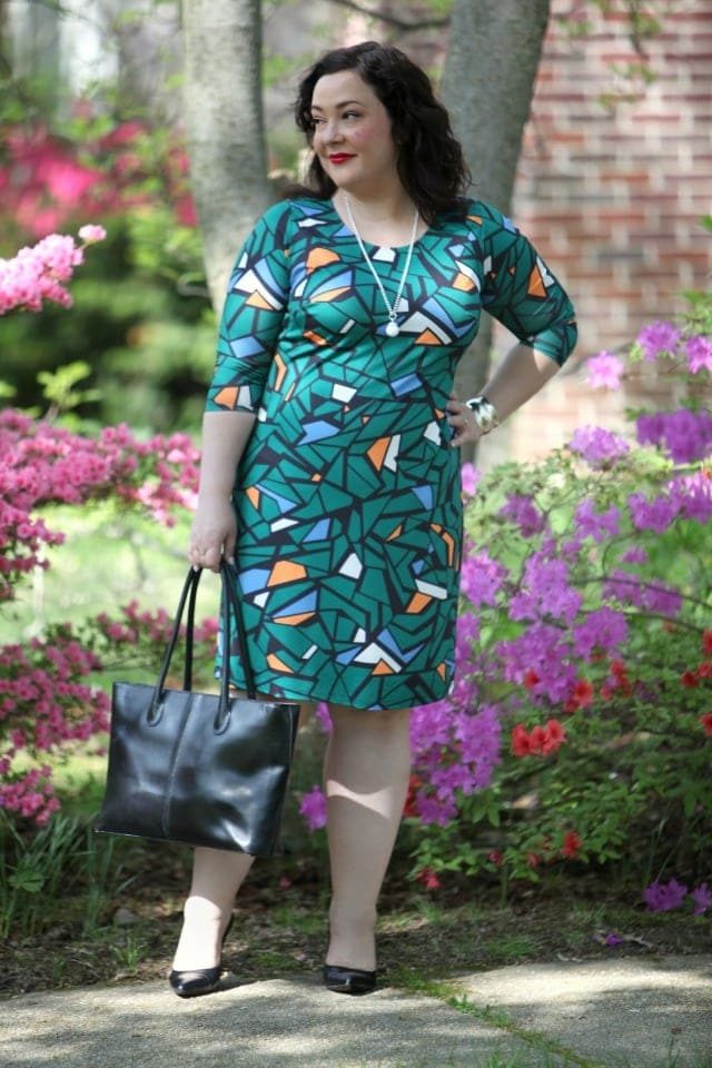 a petite size 14 woman in a green mosaic print bracelet sleeve length sheath, wearing black pumps and holding a black leather tote bag