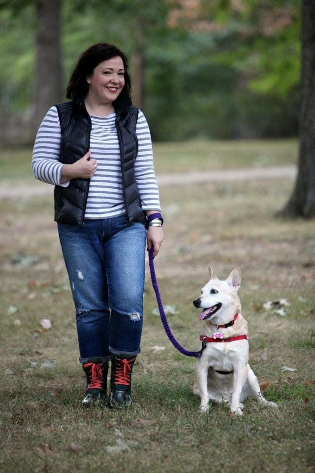 woman in jeans, a striped shirt, and black vest walking a midsized blonde dog on a leash