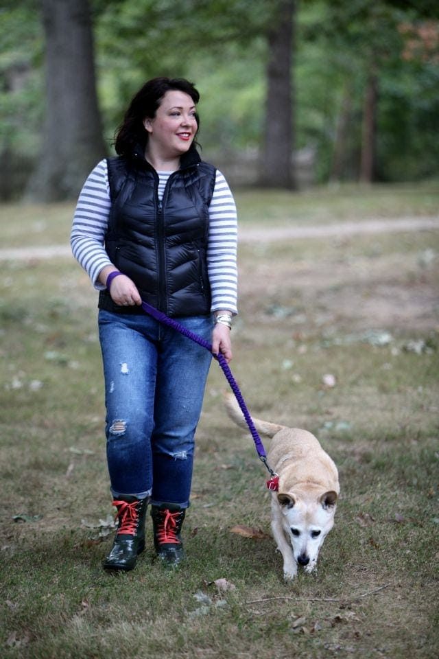 woman in jeans, a striped shirt, and black vest walking a midsized blonde dog on a leash