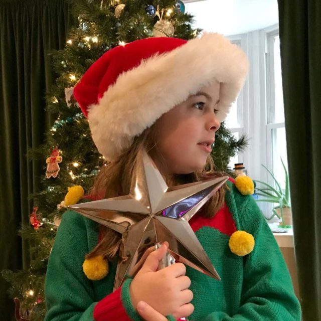 Picture of an elementary school aged girl in a Santa hat and Christmas sweater, standing in front of a Christmas tree, holding a silver Pottery Barn tree star