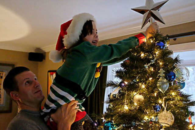 Man holding up a child as she places a silver Pottery Barn star tree topper on top of a Christmas Tree
