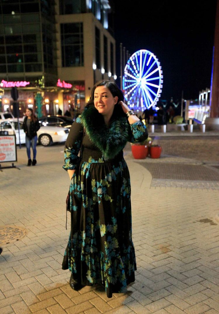 Woman in a black floral maxi dress and dark green faux fur collar standing on a street. It is nighttime and she is in National Harbor in Maryland with the Ferris wheel in the background.