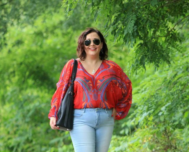 Alison Gary of Wardrobe Oxygen standing with hand on her hip, smiling. She is wearing a red printed blouse and light denim jeans with aviator sunglasses