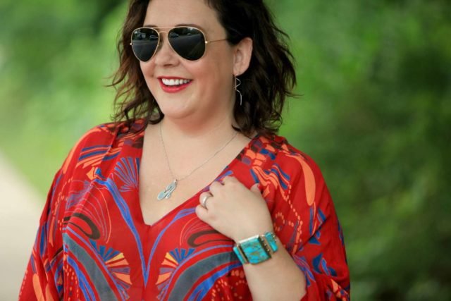 Closeup of Alison Gary smiling away from the camera. She is wearing dark aviator sunglasses, a red printed v neck blouse, and a turquoise and sterling silver cuff bracelet.