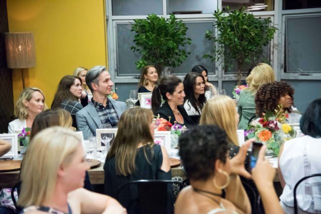 photo is of several people sitting at tables smiling and looking at someone who is off camera.