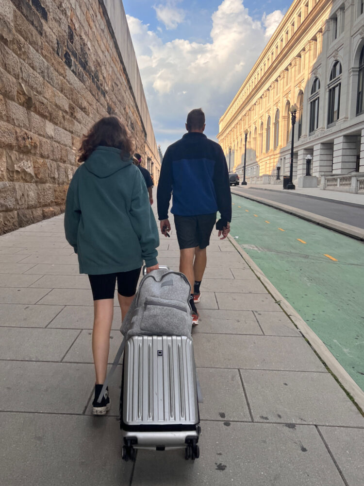 I’ve Owned the Dagne Dover Dakota Backpack for Over 5 Years: Here’s My Honest Review 1 A teen and her father walking into a train station; the teen is pulling a rolling suitcase with a gray Dagne Dover Dakota backpack on top secured by its luggage sleeve. Dagne Dover Dakota backpack review by Wardrobe Oxygen