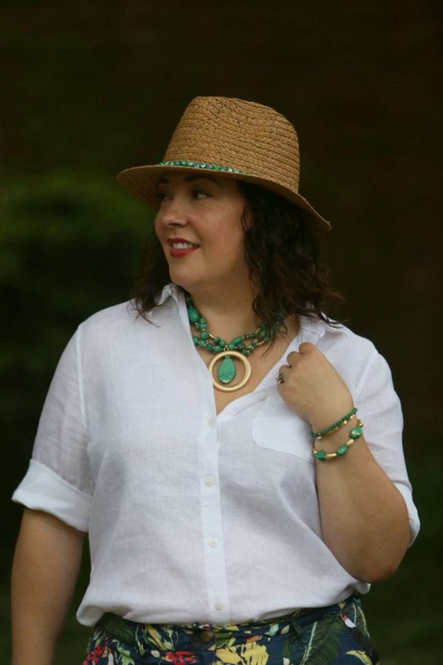 Woman with brown shoulder length wavy hair, wearing a straw fedora and turquoise jewelry with a white Chico's wrinkle free linen shirt. She os looking away from the camera and smiling.