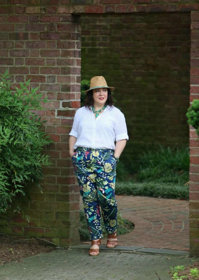 A woman in a straw fedora, white Chico's Wrinkle Free lLnen shirt tucked into floral print chinos, and tan sandals is walking through a doorway in a brick wall, entering a garden.