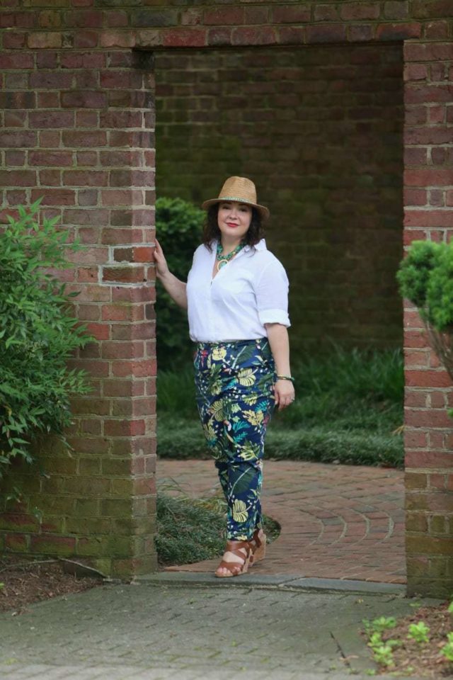 A woman in a straw fedora, white Chico's wrinkle free linen shirt tucked into floral print chinos, and tan sandals is walking through a doorway in a brick wall, entering a garden.