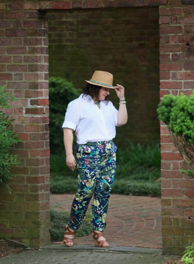 A woman in a straw fedora, white Chico's wrinkle free linen shirt tucked into floral print chinos, and tan sandals is standing in a doorway in a brick wall, entering a garden. She is holding the brim of her hat and looking down.