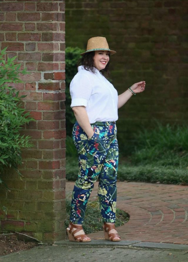 A woman in a straw fedora, white Chico's wrinkle free linen shirt tucked into floral print chinos, and tan sandals is walking through a doorway in a brick wall, entering a garden.