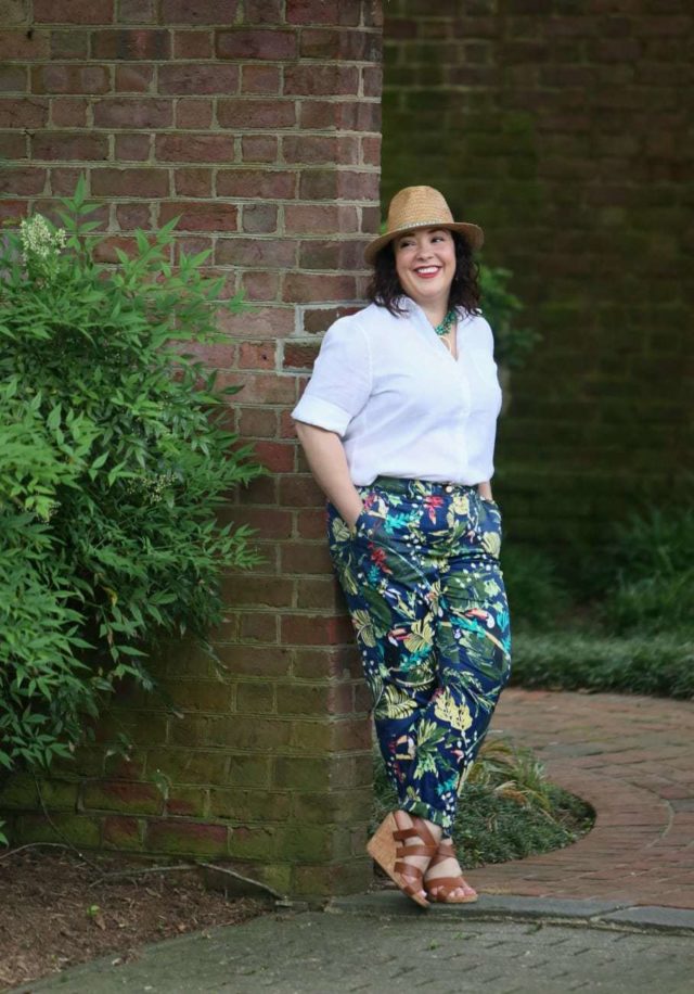 A woman in a straw fedora, white Chico's wrinkle free linen shirt tucked into floral print chinos, and tan sandals is leaning against a doorway in a brick wall, entering a garden.
