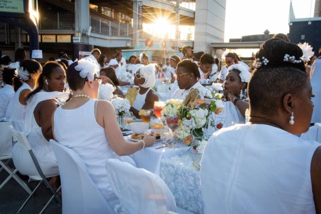 People in white sitting at long tables enjoying food at Dîner en Blanc DC as the sun sets