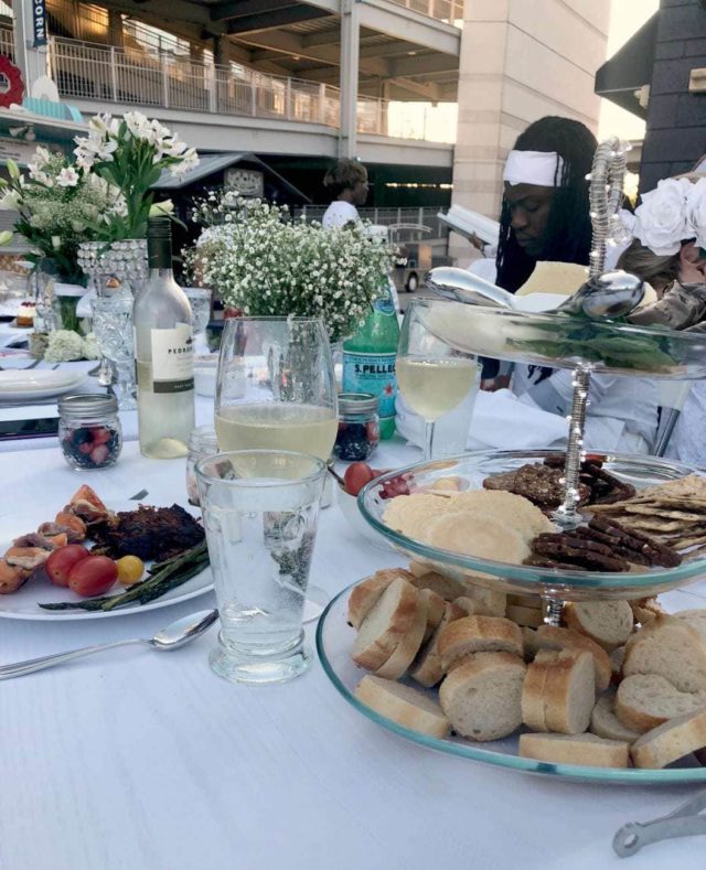 Photo of a table display at Dîner en Blanc with a glas three-tier serving dish holding food and shatter resistant wine glasses for Dîner en Blanc