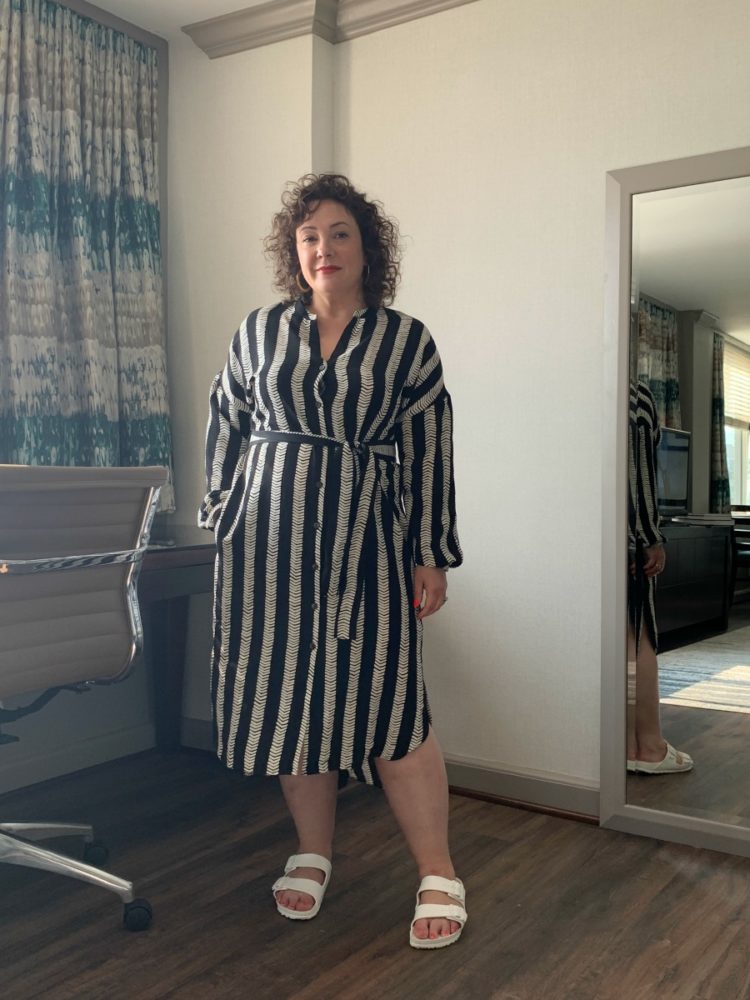 A woman wearing a black and white vertically striped belted shirtdress that hits middle of the calf length, one hand in pocket. This is for a DUBGEE by Whoopi review of the clothing line