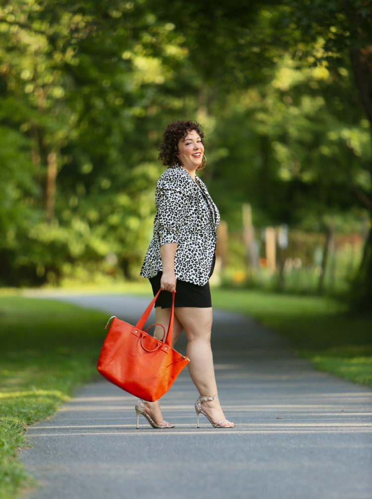 Alison wearing a leopard print blazer with a black tee and black shorts carrying an orange Clare V tote