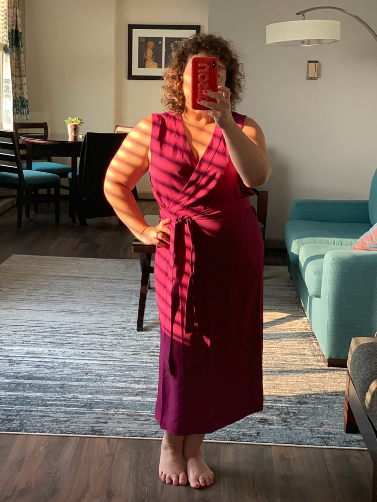 A woman standing in front of a mirror taking a selfie, wearing a magenta sleeveless wrap dress that hits mid-calf in length.