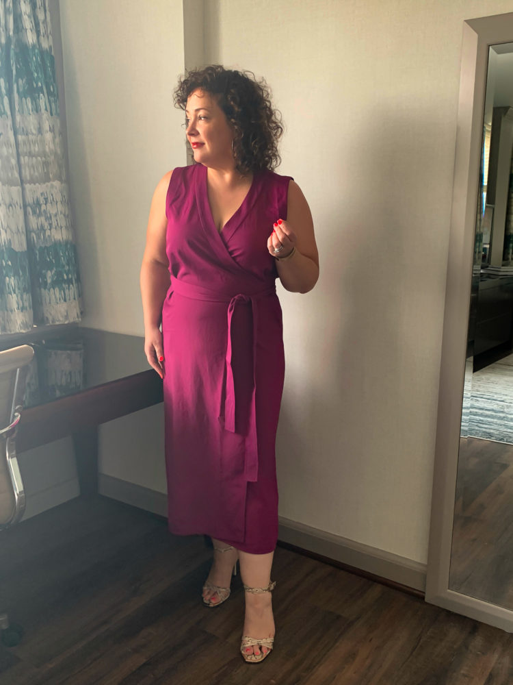 A woman in a magenta wrap dress standing in the corner of a room looking out the window