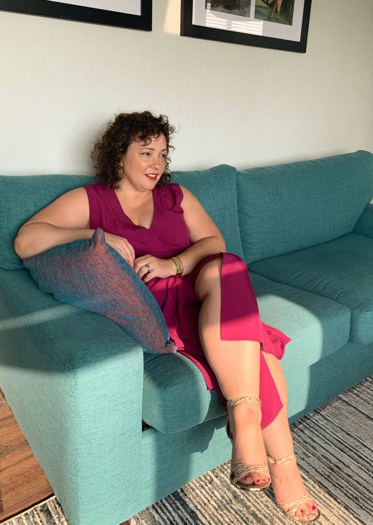 Alison Gary of Wardrobe Oxygen wearing a magenta colored wrap dress, sitting on a turquoise couch, her legs crossed.