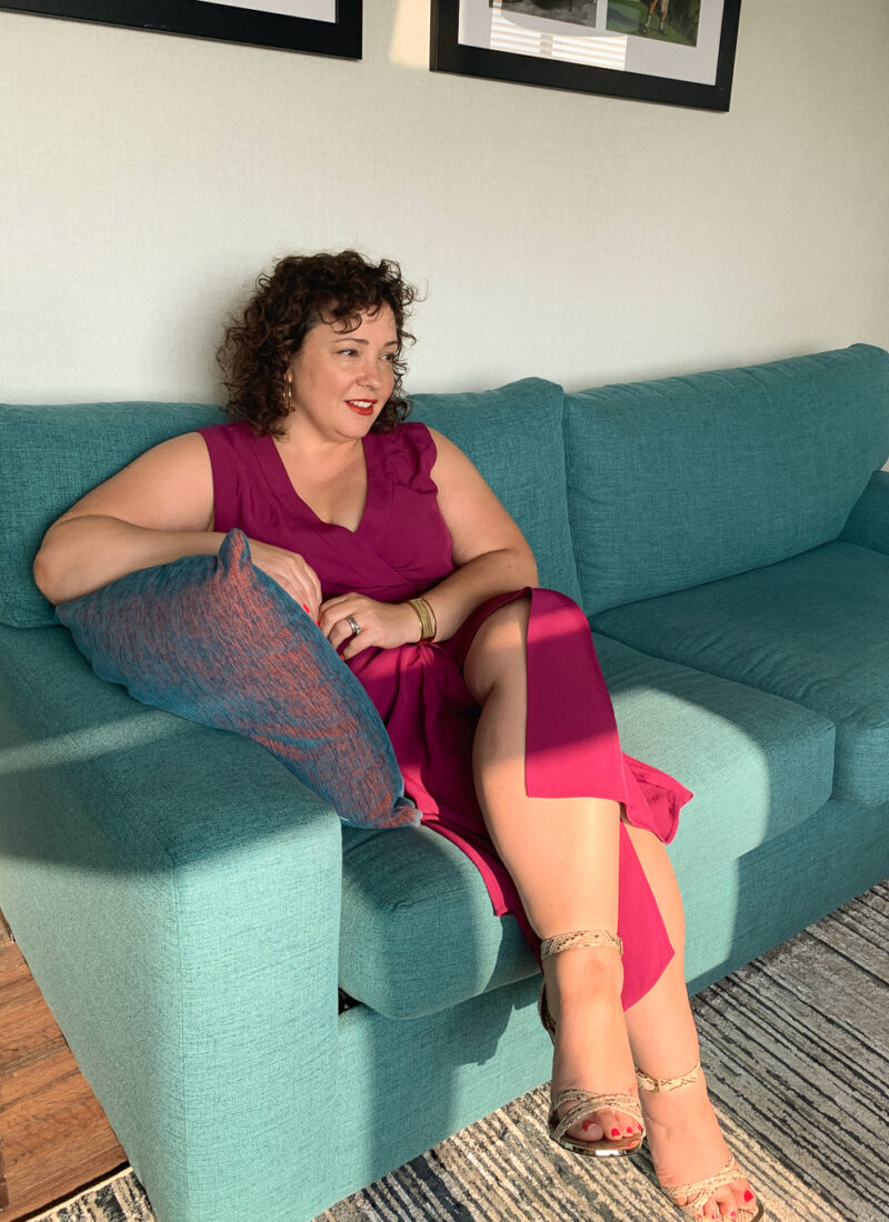 Alison Gary of Wardrobe Oxygen wearing a magenta colored wrap dress, sitting on a turquoise couch, her legs crossed.