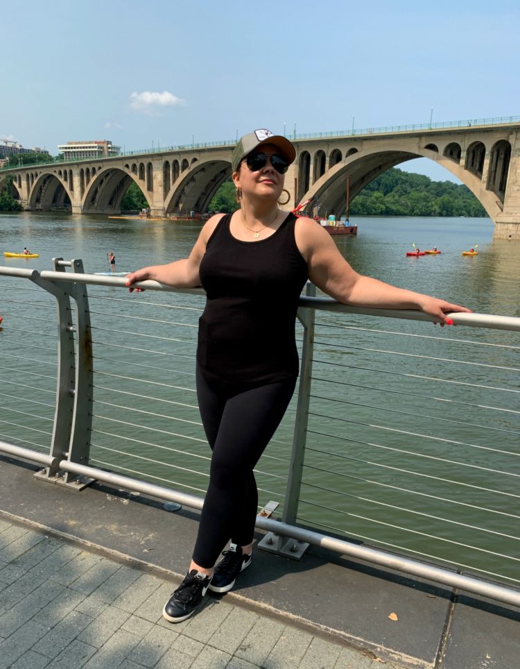 Image of a woman in a black tank and black leggings leaning against a railing. There is the Key Bridge behind her and water where there are several people in kayaks and canoes. The sky is blue.