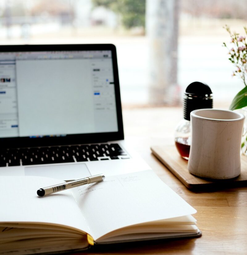 Open notebook with pen in front of open laptop with cup of coffee all on a desk in front of an open window