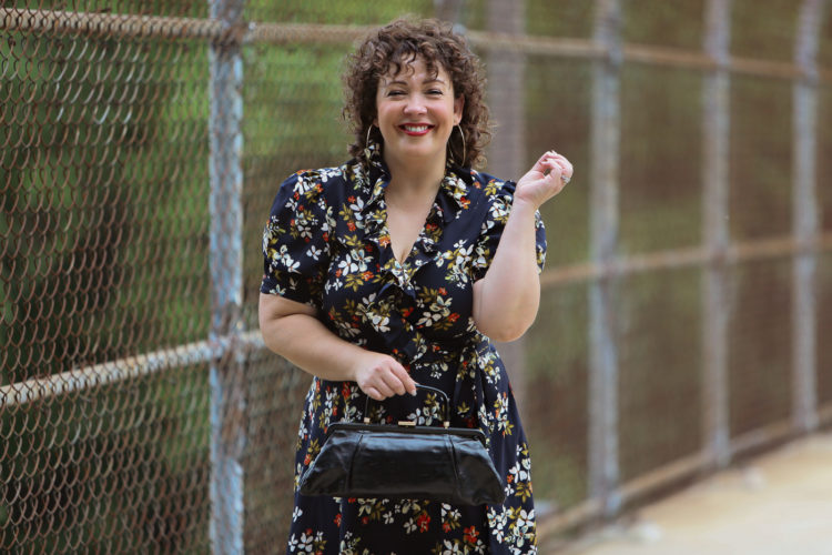 Alison Gary of Wardrobe Oxygen in a navy floral wrap dress holding a black leather vintage-inspired frame bag and smiling at the camera