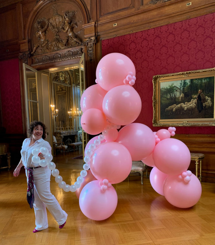 woman smiling and holding onto a chain made of balloons that is attached to a 12 foot tall pink poodle also made of balloons