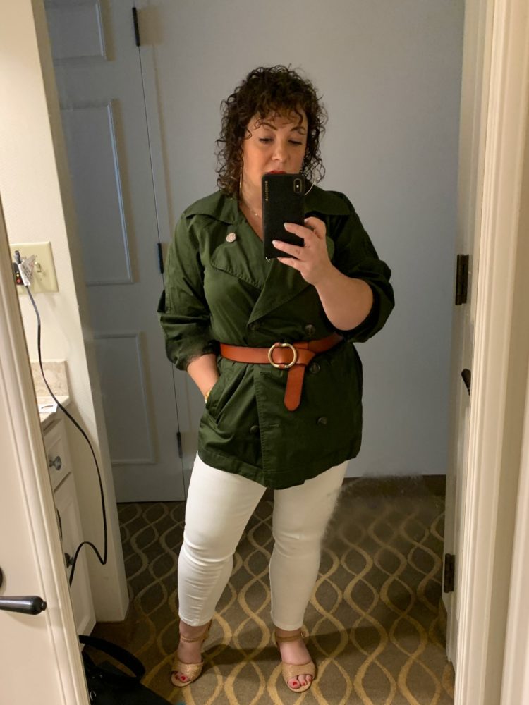 woman taking mirror selfie while wearing olive jacket and white jeans