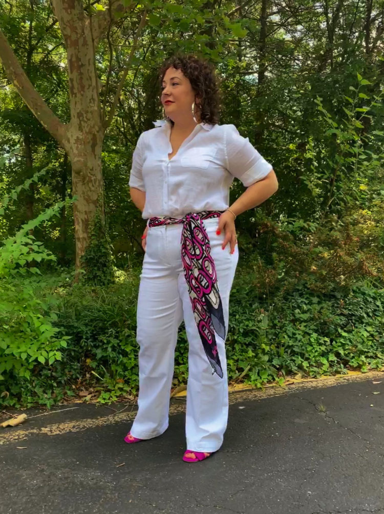 woman in white linen shirt and white wide leg jeans with a magenta and navy scarf as a belt. Her hands are on her hips and she's looking off into the distance.