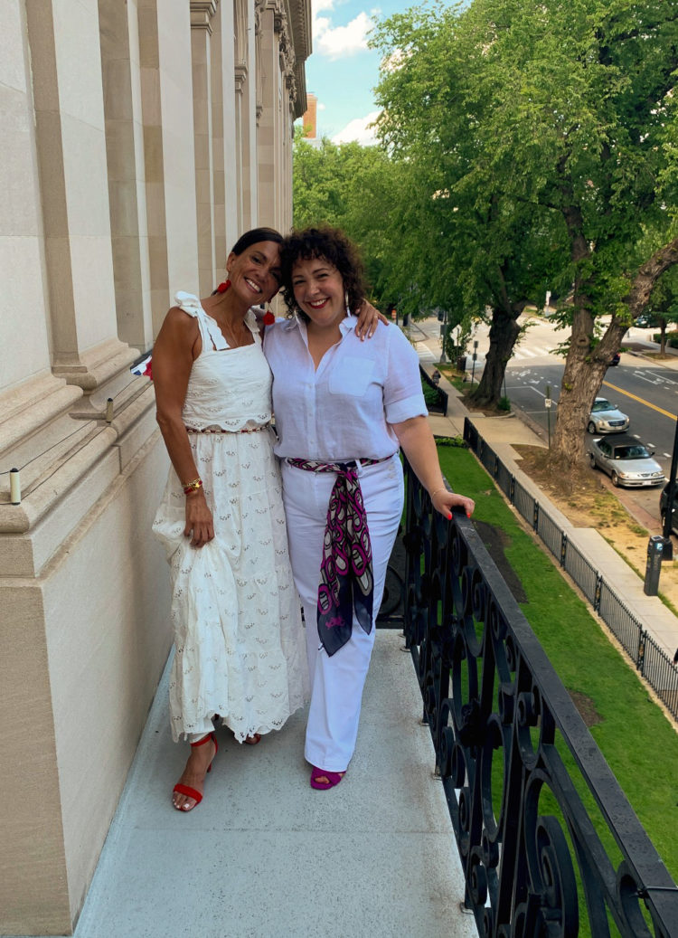 two brunette women with their arms around one another's shoulders. They are both dressed in summery white clothing and smiling.