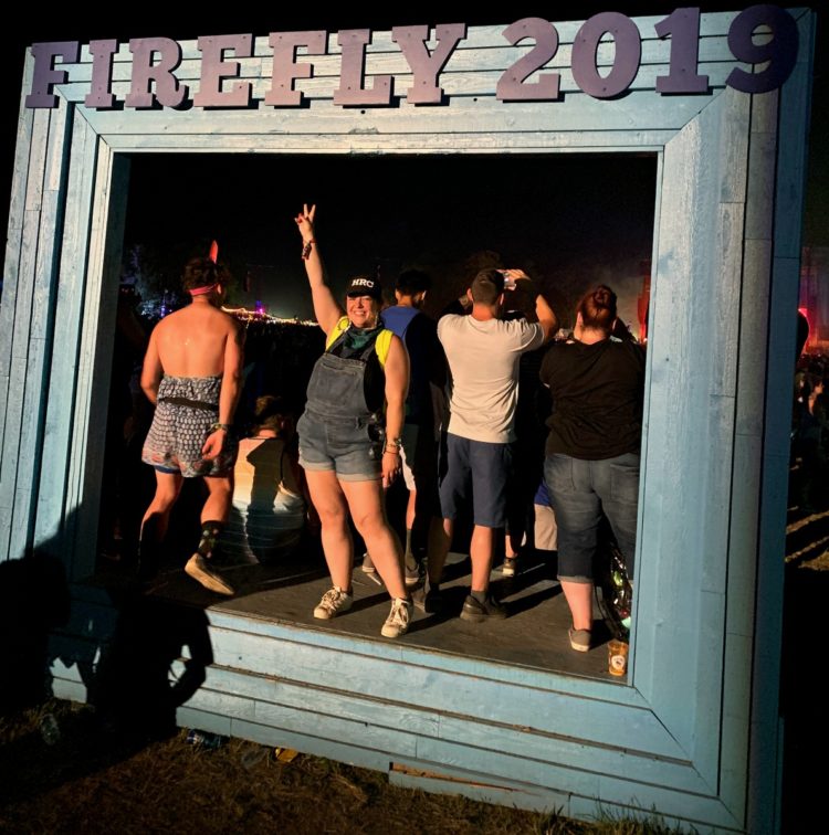 woman in shortalls giving the peace sign while standing on a stage that says FIREFLY 2019