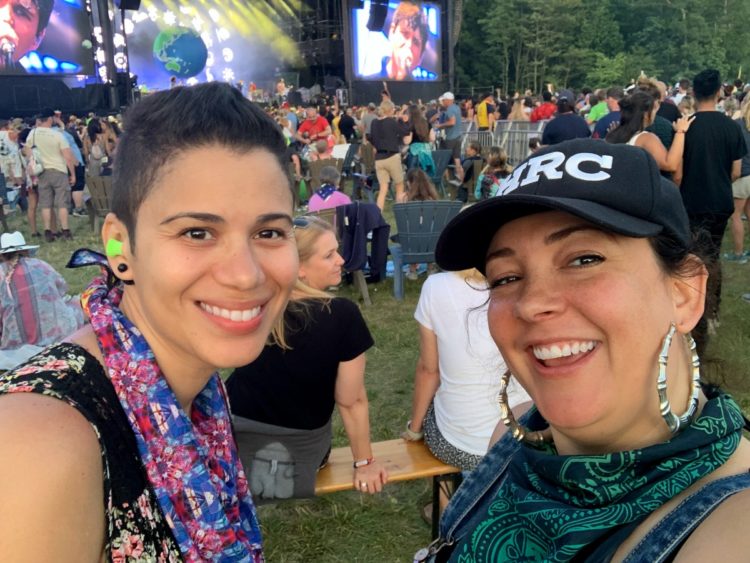 two women in bandanas smiling at the camera with Vampire Weekend on stage behind them