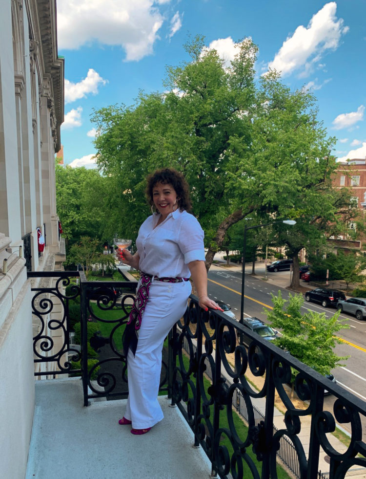 woman in all white leaning against a balcony railing in washington DC, holding a glass of champagne and smiling.