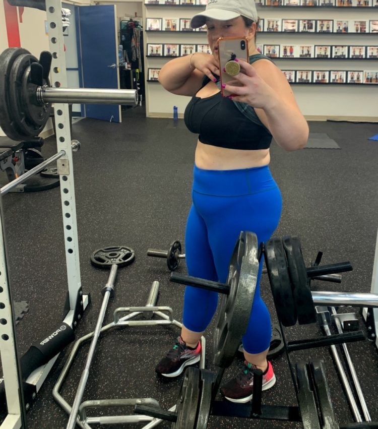 woman taking mirror selfie in blue leggings and lifting her shirt to show her sports bra