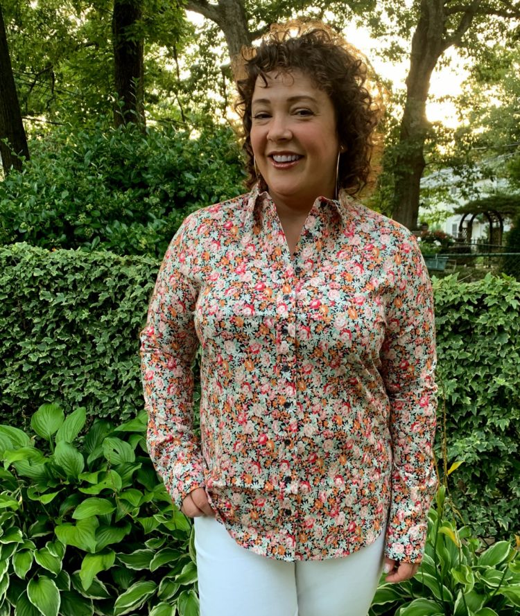 woman in pink and orange floral button front long sleeved shirt smiling at the camera with one hand in the front pocket of her white jeans