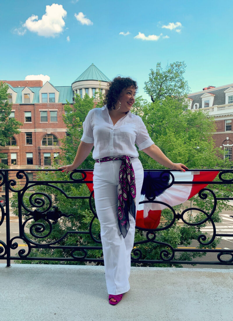 woman in white linen shirt tucked into white high rise wide leg denim trousers with a navy and magenta chiffon scarf tied around her waist as a belt. She is standing on a balcony overlooking Dupont Circle in DC and smiling.