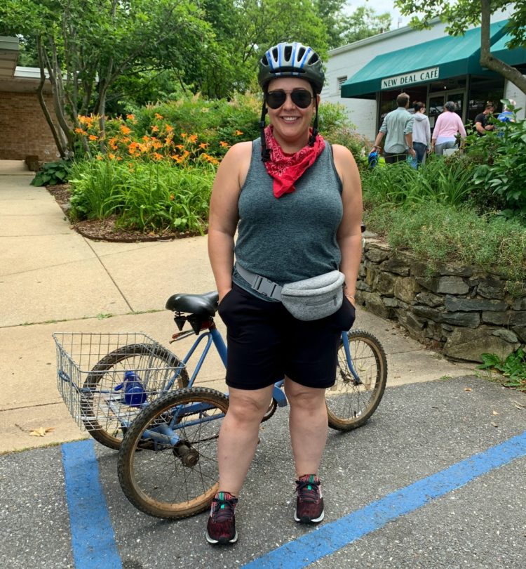 woman with hands in her pockets smiling white standing in front of an adult tricycle