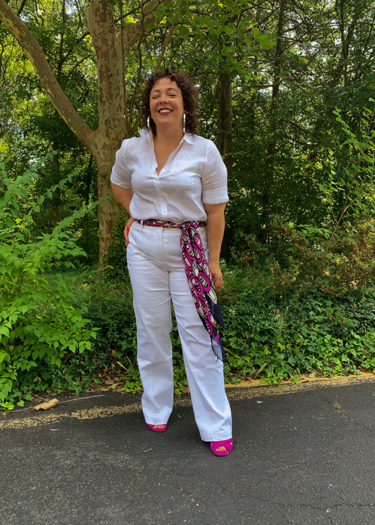 Woman in white linen shirt and white jeans with a colorful scarf tied as a belt smiling with hand on hip