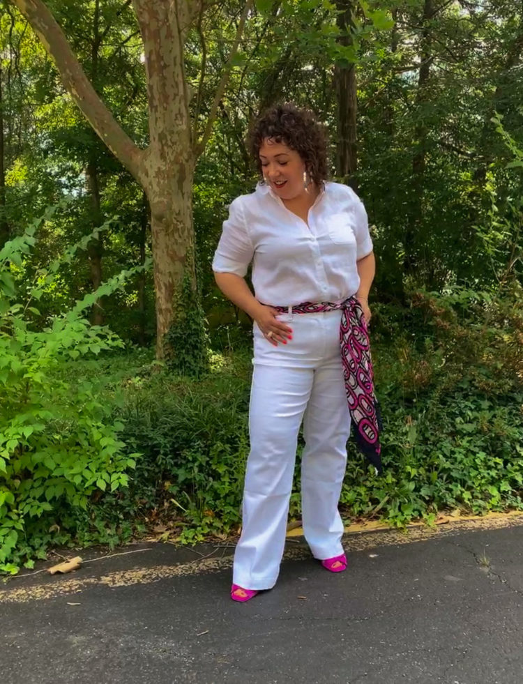 Woman in white linen shirt and white wide leg jeans standing in front of a forest looking down at her shoes.