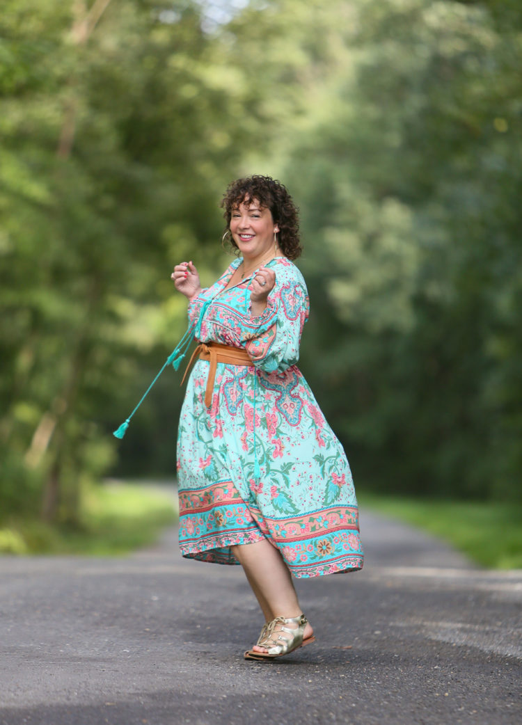 woman in a green floral dress and tan belt spinning on a gravel road in the woods