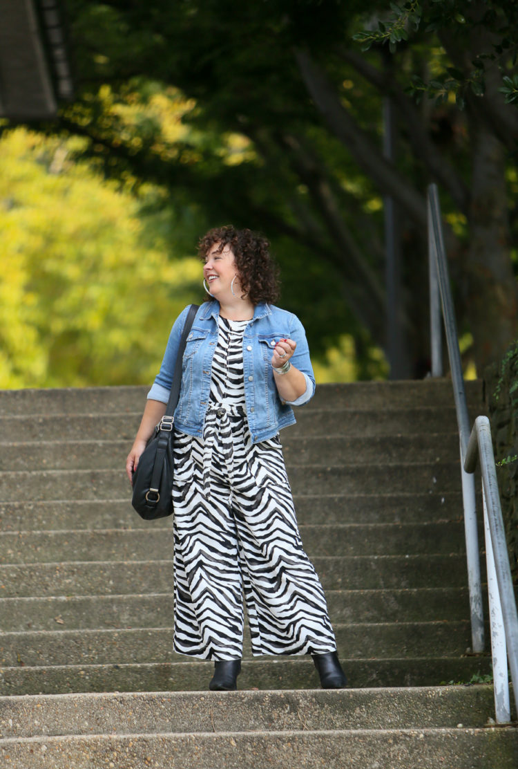 Styling a summer jumpsuit for fall - tips on how to do this successfully by Wardrobe Oxygen who is wearing a zebra print Banana Republic jumpsuit with a denim jacket and black ankle booties by Clark's and carrying a Rough & Tumble handbag in black leather.